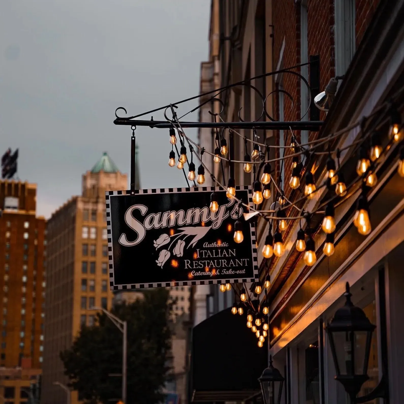 Sammy's hanging sign over 3rd Street at dusk, strung with warm lights